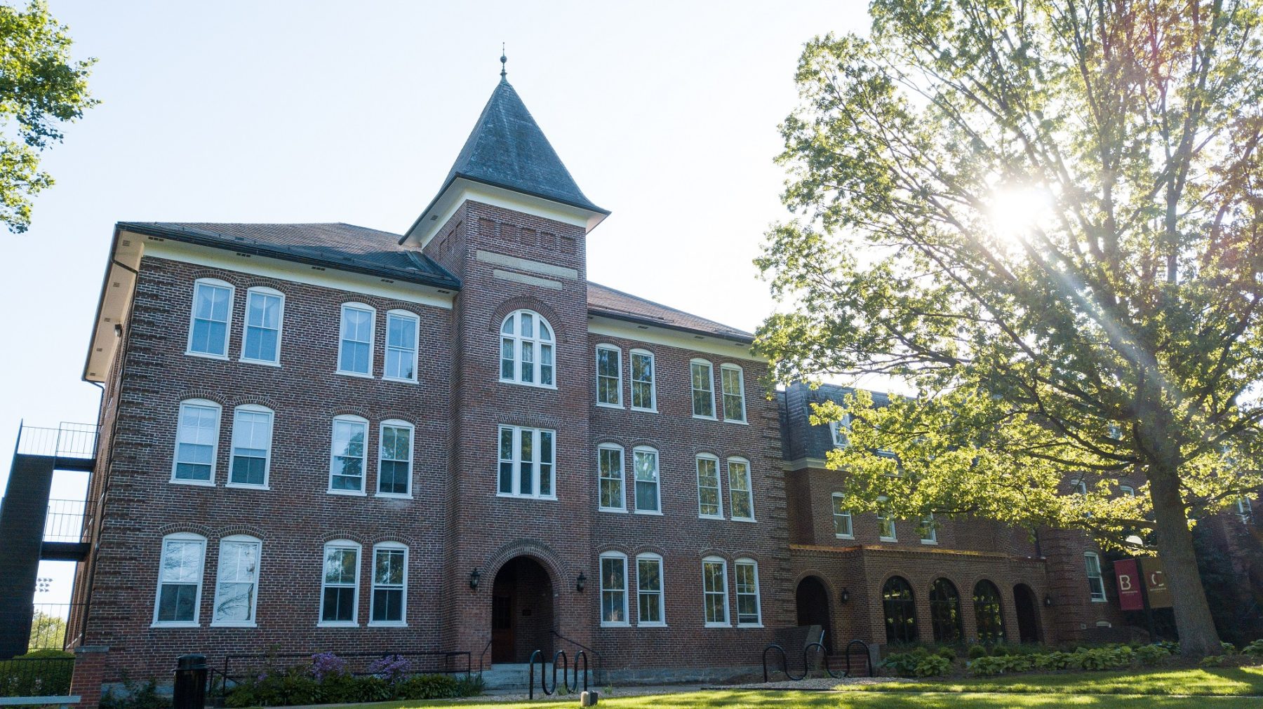 A brick building on Bridgewater College's campus in May of 2020 with a tree blooming in the front and the sun peeking through its branches