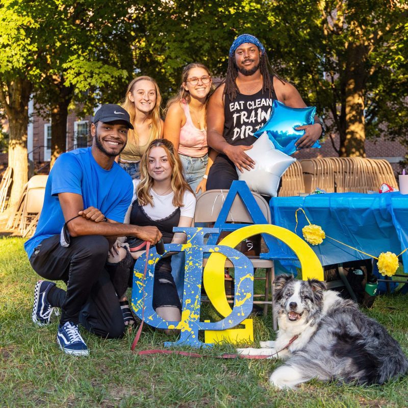 Group of students posing for photo with a sign and dog in front