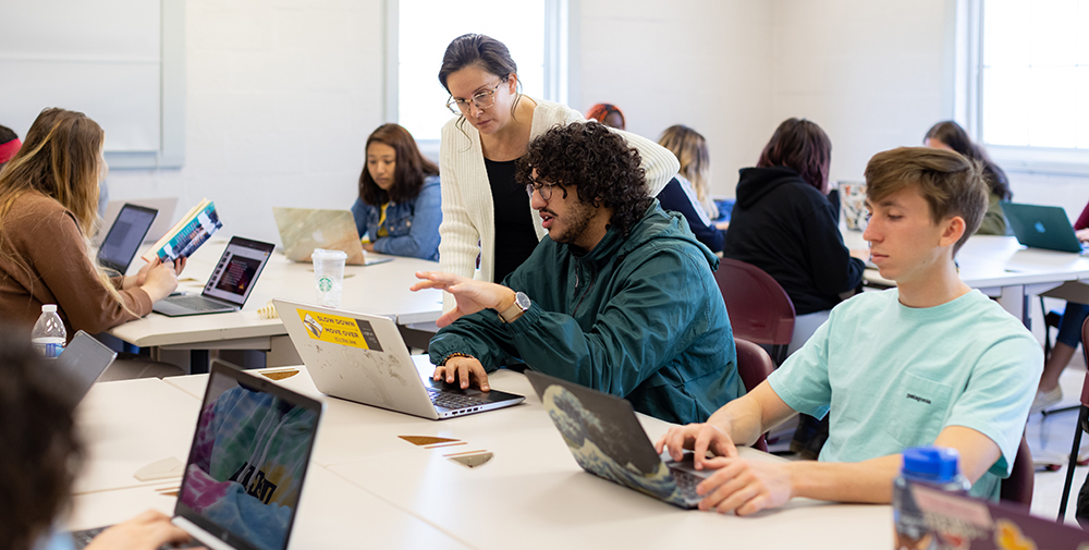 Professor helping a student while looking at a laptop surrounded by other student in the classroom