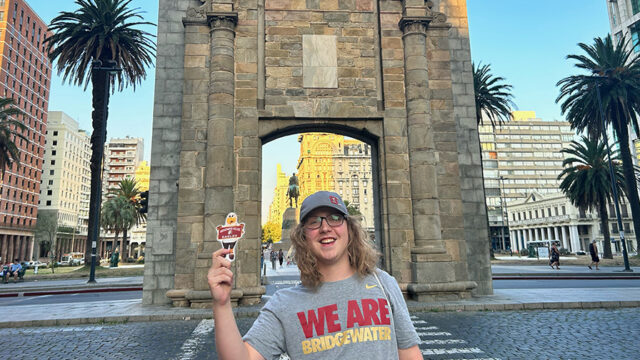 Student standing in front of the Gateway to Citadel in Uruguay in a Bridgewater College tshirt and holding flat Ernie