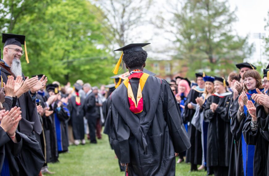 A student in a graduation cap and gown walks through an echelon of professors also in graduation regalia