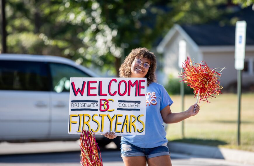 A female student holding a sign that says welcome B-C first years. In her other hand is a pompom